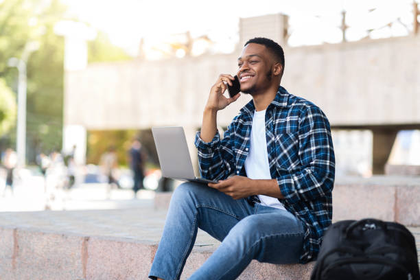 istock Black man using laptop, sitting in the city with smartphone 1264235458