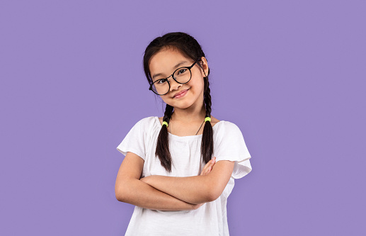Little Chinese Girl Wearing Eyeglasses Posing Crossing Hands Standing Over Pastel Purple Background, Looking At Camera. Studio Shot