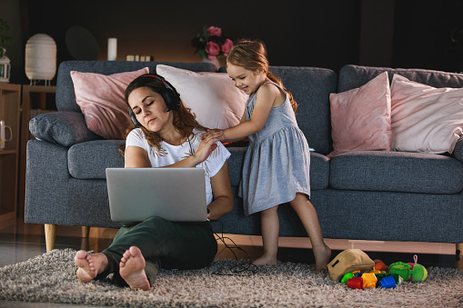 Front view of a focused entrepreneur using her laptop computer to work remotely during isolation while her little daughter comes to plays with her.