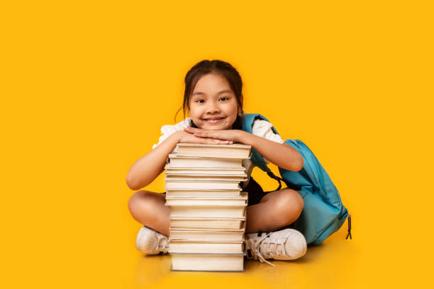 istock Asian Schoolgirl Sitting At Book Stack On Yellow Studio Background 1264243383