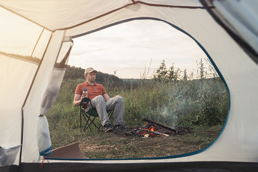Young man sits on a chair near a bonfire against the backdrop of nature. View through the tent. Evening time, dramatic sky. Concept of local travel, tourism and camping. Copy space.