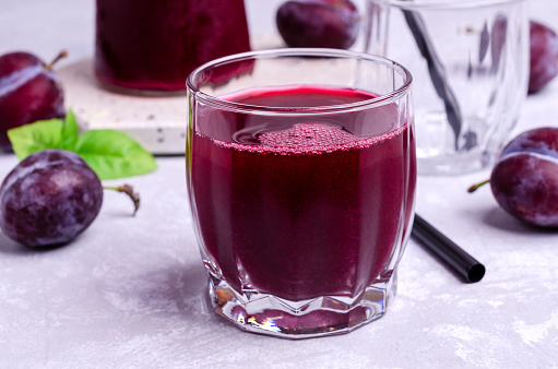 Red plum juice in glass on a gray stone background with fresh fruit. Selective focus.