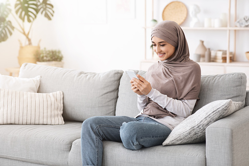 Modern Muslim Girl Using Smartphone At Home While Relaxing On Couch In Living Room, Texting With Friends Or Browsing Social Networks