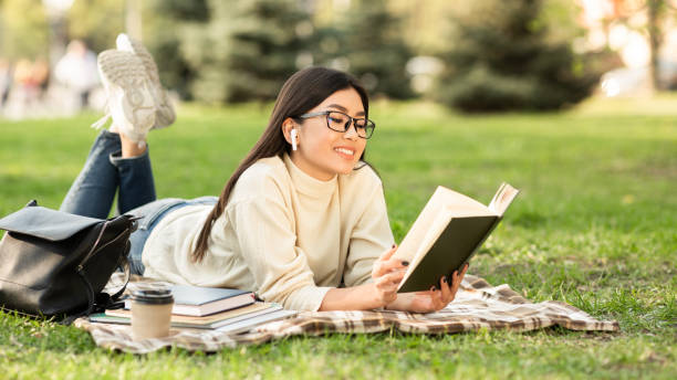 girl reading a novel lying on the grass - woman with glasses reading a book imagens e fotografias de stock