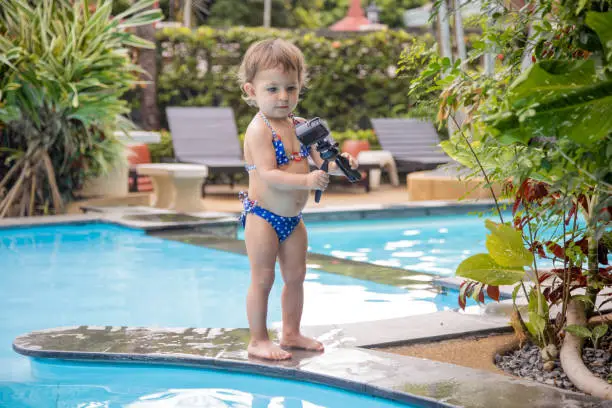 Photo of young blogger. cute toddler girl in a swimsuit stand by the pool and hold action camera.