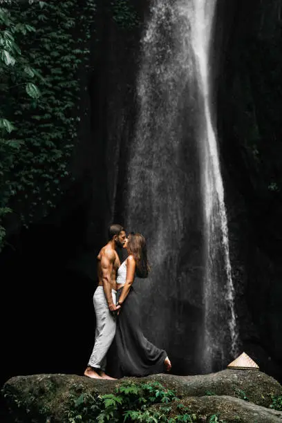Photo of Beautiful couple at a waterfall in Indonesia. A couple in love travels around the island of Bali. A man and a woman kiss at the waterfall. Travel to Asia. Honeymoon trip. Tourists at the waterfall