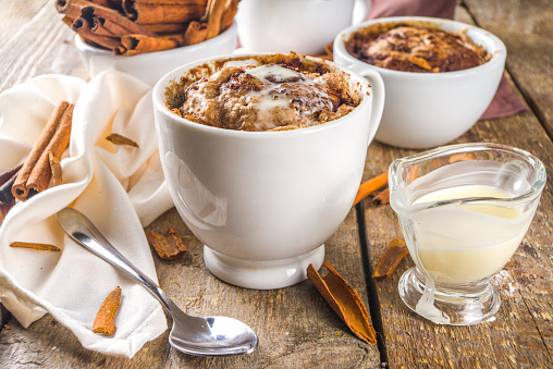 Cinnabon cake in mug. Fast simple microwave dessert idea, background for recipe. Cinnamon roll mugcake, with sugar and cream cheese topping, in different mugs, with cinnamon sticks on wooden background