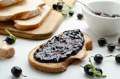 Process of spreading of sweet delicious jam on the slice of bread.Closeup of spoon, blackcurrant jam, berries, bread on the white table.Tasty jam sandwich