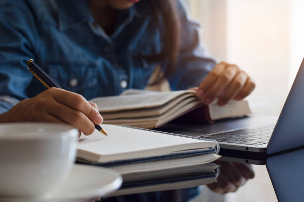 Female student Casual young female student handwriting information on diary notebook while reading book and work on laptop computer with white cup of coffee on the table at home. learning stock pictures, royalty-free photos & images