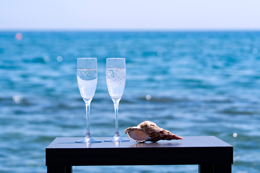 Two glasses with sparkling wine and seashell in front of blue sea at sunset
