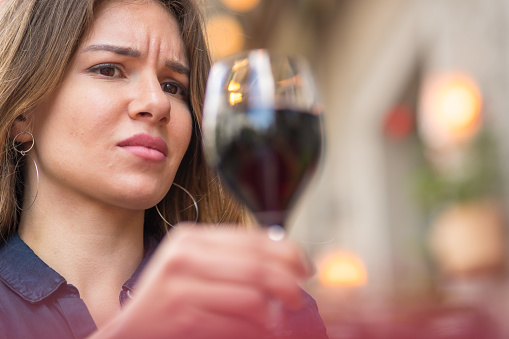 Close up of a woman with a disgusted expression looking at a glass red wine she is holding, as to refuse alcohol.