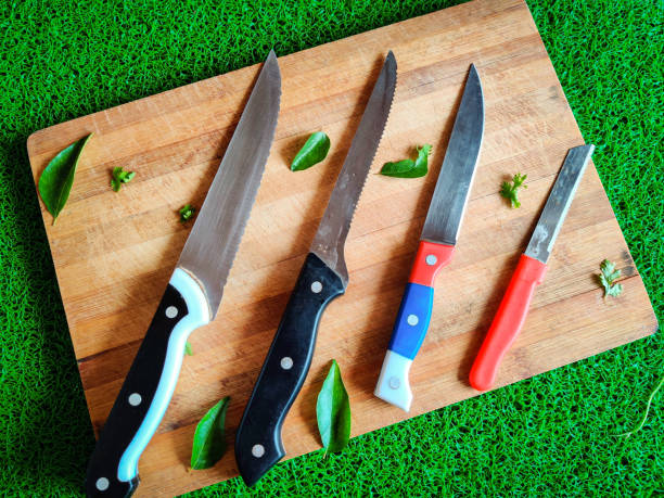 Four colored different sizes of knives kept on chopping board. With curry leaves and coriander leaves spread around