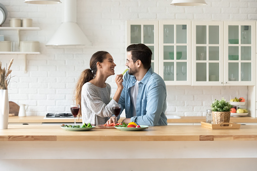 Young loving couple tenants enjoy romantic date cooking together drinking wine in home kitchen, happy millennial husband and wife celebrate wedding anniversary, have family weekend celebration