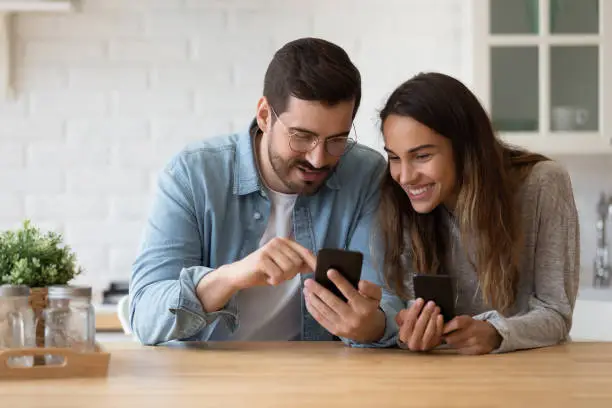 Smiling millennial couple sit at table in kitchen have fun using modern smartphone devices together, happy young husband and wife laugh relax at home browsing application on cellphone gadgets