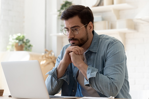 Pensive young Caucasian man in glasses sit at desk look at laptop screen thinking pondering, thoughtful millennial male in spectacles work at computer consider ideas solving problems at home workplace