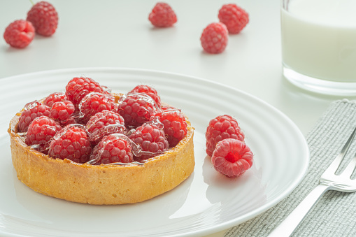 Raspberry tartlets served on a transparent plate with brown sugar. Glass of milk in the sun on a white background.