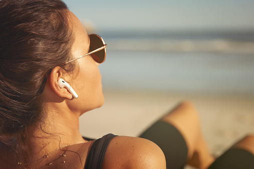 Young woman is sitting on the beach listening to music with air-pods after a workout.  Ocean in background