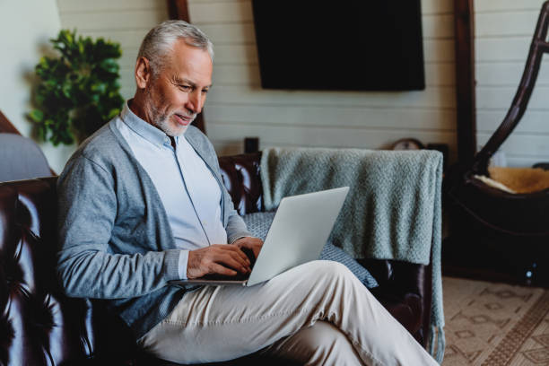 istock Handsome old man using laptop and smiling while sitting on couch at home 1264328326