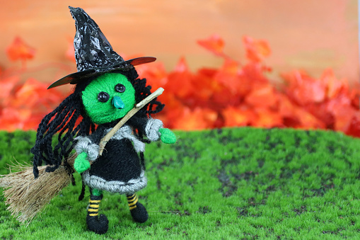 Stock photo showing close-up view of string model Halloween witch with broomstick pictured in autumnal country scene with green grass, orange hedgerow and sunset sky.