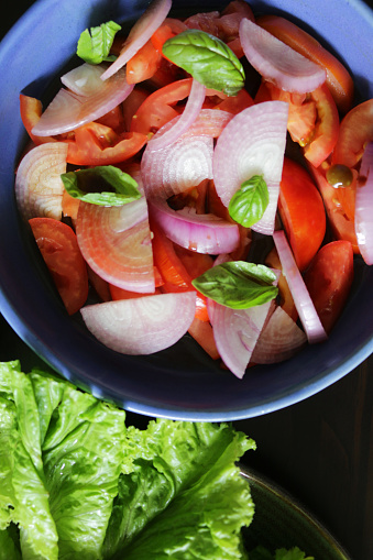 Stock photo showing elevated view of blue bowl containing tomato and onion salad. Slices of tomato and red onion marinated in vinegar and oil, garnished with basil leaves.