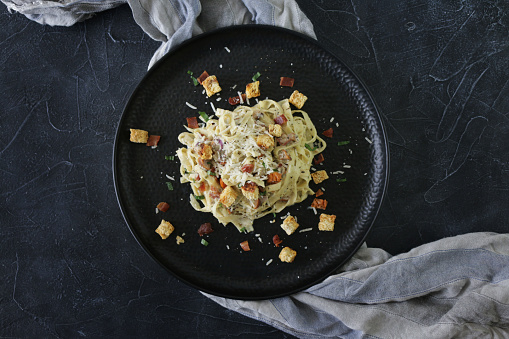 Stock photo showing an elevated view of a black dimpled plate with a portion of homemade fettuccine carbonara, using a traditional Italian recipe. The pasta is mixed with eggs, herbs, crispy bacon lardons and pancetta, prosciutto slices, grated Italian Parmesan cheese and garlic.