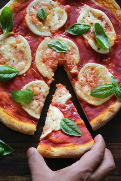Stock photo showing elevated view of a Margherita pizza topped with a rich tomato sauce, melted circles of buffalo mozzarella and garnished with fresh basil leaves, against a round wooden chopping board background. imagen de primer plano de persona irreconocible sosteniendo una rebanada triangular de pizza margherita casera recién horneada, círculos de queso mozzarella de búfalo dorado, rica salsa marinara de tomate y hojas frescas de albahaca sobre fondo redondo