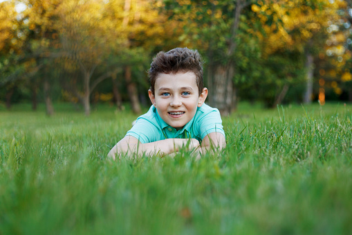 A smiling young boy rolls in the green grass, enjoys the park, looks at the camera on a sunny summer day.