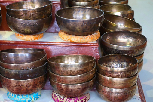 Stock photo showing a stack of copper Tibetan singing bowls, displayed piled on wooden shelves for sale. These bowls can be used in sound therapy either by being struck by a wooden striker or the striker being run around the rim.
