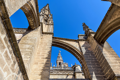 Seville, Andalusia, Spain - October 9, 2019: A close-up low-angle view of the pinnacles and flying buttresses on the roof of Seville Cathedral, with top of La Giralda tower in background.