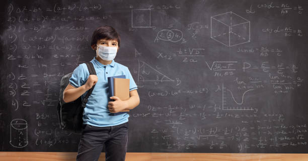 scolaro che indossa una maschera protettiva per il viso e si trova di fronte a una lavagna della scuola - little boys measuring expressing positivity intelligence foto e immagini stock