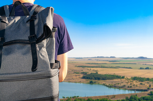 Close-up backpack at back of men traveler looking at the horizon under a blue sky.