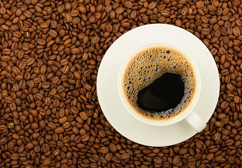 Close up white cup on saucer full of black coffee over background of roasted coffee beans, elevated top view, directly above