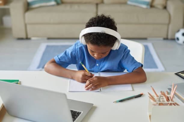 istock Focused African-American schoolboy in wireless headphones making notes in workbook while listening to online lecture 1264348861