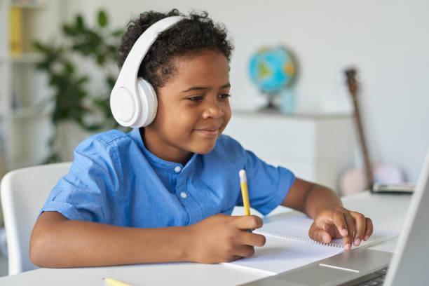 istock Content inquisitive Black boy in wireless headphones sitting at desk and using laptop while working with tutor 1264348863