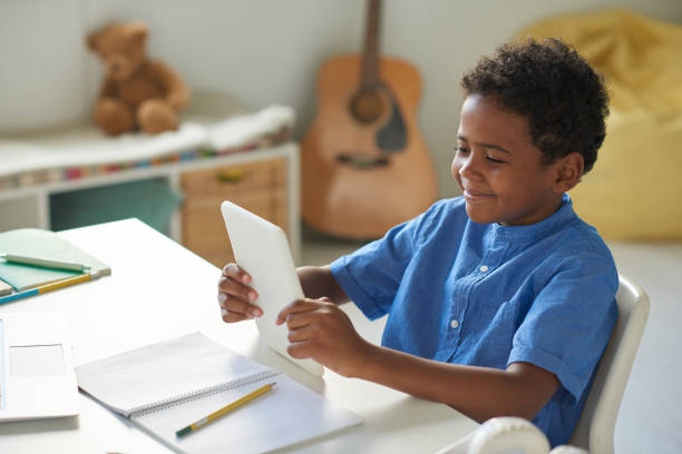 istock Smiling cute black boy in blue shirt sitting at desk with workbook and using learning app on tablet 1264348899