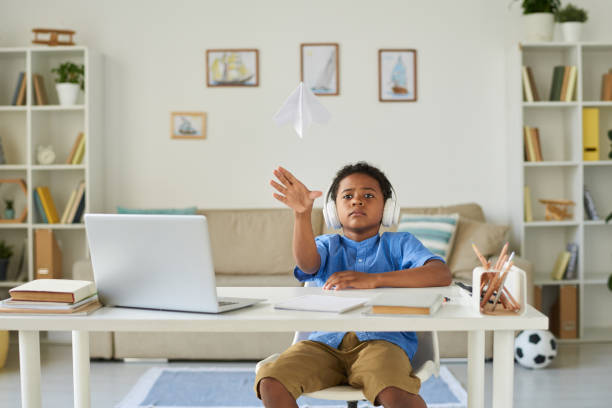 istock Lazy African-American boy in wireless headphones throwing paper plane while procrastinating instead of learning 1264348900