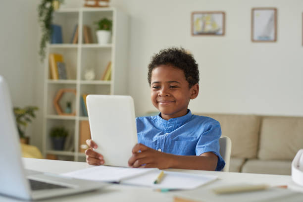 istock Content cute Afro-American boy sitting at table in living room and using tablet while communicating via video conferencing app 1264348902