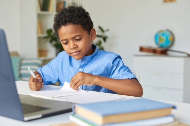 istock Curious black schoolboy with curly hair sitting at table and checking notes in workbook while doing homework 1264348967