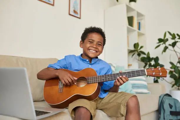 Photo of Portrait of cheerful adolescent black boy sitting on sofa in living room and playing guitar according to video lesson