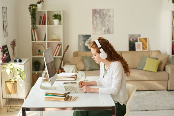 Concentrated redhead student girl in wireless headphones sitting at table and writing down thesis while listening to online lecture Writing down thesis while listening to online lecture dissertation stock pictures, royalty-free photos & images