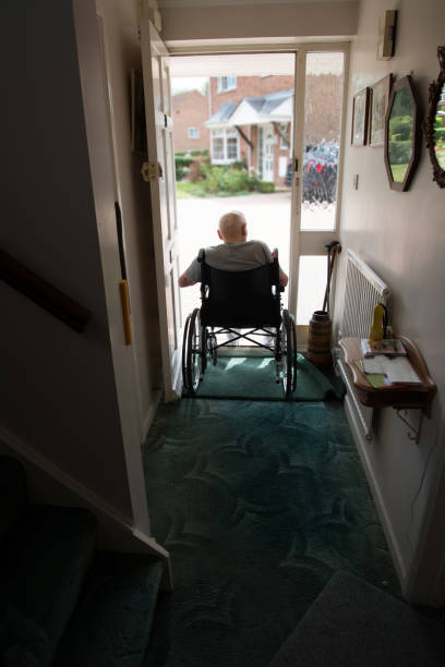 vecchio in sedia a rotelle che guarda fuori dalla porta aperta della sua casa. hampshire, regno unito. - housebound foto e immagini stock