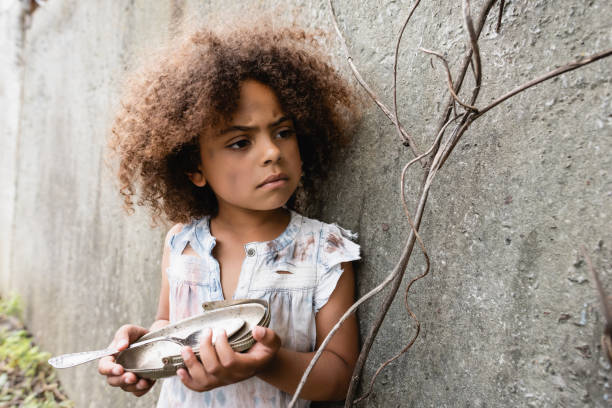 selektiver fokus von armen afrikanischen amerikanischen kind hält schmutzige metallplatte und löffel in der nähe von betonwand auf der städtischen straße - street child stock-fotos und bilder
