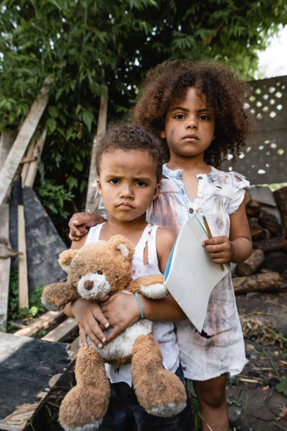 arme afrikanische amerikanische kind hält leeres papier und bleistift, während in der nähe von traurigen bruder mit teddybär stehen - street child stock-fotos und bilder
