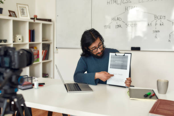 istock Young caucasian man teacher wearing eyeglasses teaching how to code HTML, sitting at his workplace and giving online lesson. Recording video blog 1264356043