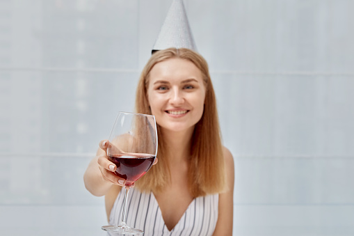 Birthday celebration, holding a glass with red wine in his hand. Light background. Woman in a festive cap.