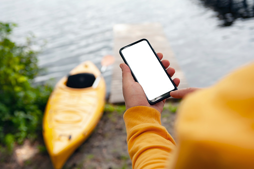 The tourist holds a phone in his hands. Mock up smartphone close-up on the background of a kayak and a lake. Concept on the topic of tourism and recreation