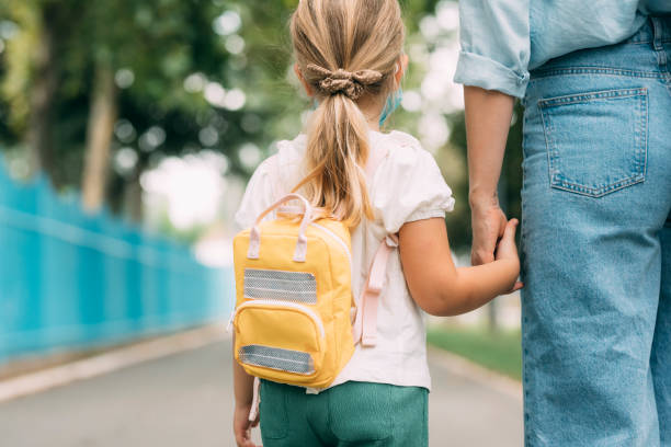 cute blonde girl wearing a protective mask being taken to school by her mom/ a babysitter - first day of school imagens e fotografias de stock