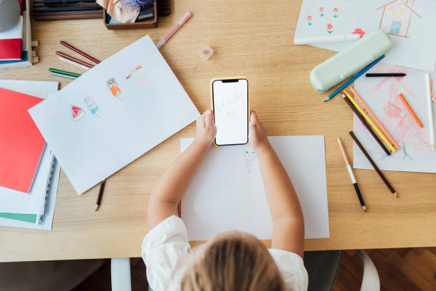 Cute Preschooler Using a Mobile Phone to Learn New Things Online, an Overhead View Homeschooling technologies: an adorable blonde girl using a smartphone to follow a drawing lesson at home. preschooler caucasian one person part of stock pictures, royalty-free photos & images