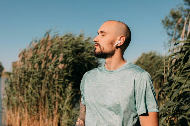 Exercising in Nature: a Relaxed Man in Sportswear Listening to Music Through his Wireless Earphones While Working Out Outdoors Staying healthy and active: a handsome Caucasian man listening to music while meditating in nature, self care concept. man yoga tattoo stock pictures, royalty-free photos & images