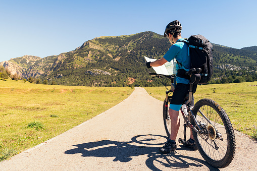 young cyclist with backpack and mountain bike standing on a road while consulting the route of his excursion on the map, concept of sport and healthy lifestyle in nature, copy space for text